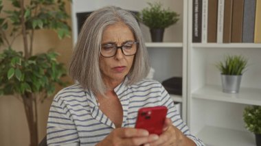 Elinde kırmızı akıllı telefon tutan ve elleri kitaplığın yanındaki ofiste görünen bir kadın, çatık kaşlı ekrana bakıyor; endişe alarmı.