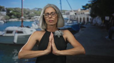 Gri saçlı ve gözlüklü bir kadın, dua ederken elleri birbirine bastırılmış, gözleri deniz kenarında bir sokakta kapalı; huzur.