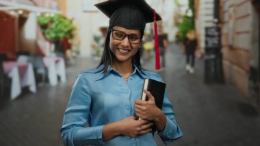 Hispanic woman in graduation attire happily holds a book on a lively restaurant terrace, expressing success and joy in an outdoor street environment.