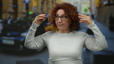 Orta yaşlı İspanyol kadın hareketli araçlar ve şehir hayatı ile çevrili, kıvırcık saçlı ve gözlüklü, günlük beyaz bir kazak giyen, işlek bir şehir caddesinde neşeli jestler yapıyor..