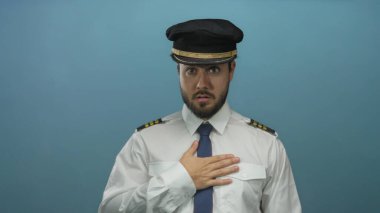 Sakallı ve üniformalı genç pilot, kafa karışıklığını ifade eden, havayolu, İspanyol ve profesyonel havacılık temalarını vurgulayan mavi bir arka plana karşı duruyor..