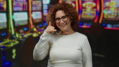 Woman making call gesture in a casino with colorful slot machines in the background, smiling and having a fun time indoors in a lively gambling environment.
