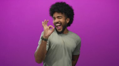 Kıvırcık saçlı bir adam canlı pembe arka planda gülümserken normal bir jest yapar. Rahat bir tişörtle ifade ve enerji dolu pozitif bir anı yakalar..