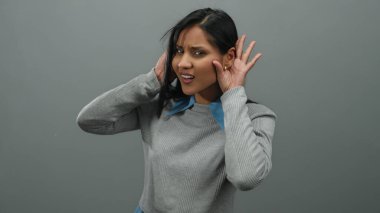 Genç İspanyol kadın, elleri kulaklarının üzerinde, gri bir arka plana karşı durarak, gündelik bir ortamda merak ve ilgi göstererek bir dinleme hareketi yapıyor..