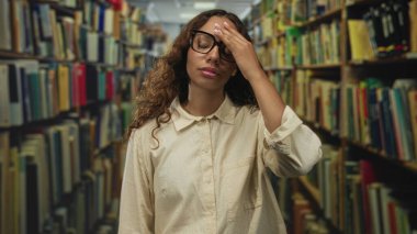 Kütüphane koridorunda kitapların arasında eli alnında gözlük takan genç bir kadın yorgunluk çalışıyor..