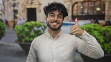 Gözlüklü ve sakallı İspanyol genç adam kalabalık bir sokakta dostça ve olumlu bir hava sergiliyor..