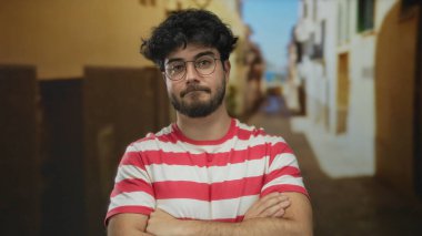 Sakallı ve gözlüklü genç adam kollarını açık bir sokakta çapraz yapıp kırmızı ve beyaz çizgili bir gömlek giyerek rahat ve rahat bir hava veriyor..