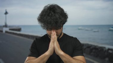 Sakallı genç adam deniz kenarında dua ediyor. Okyanusun sakin zemininde, bulutlu bir gökyüzünün altında bağlılığını gösteriyor..