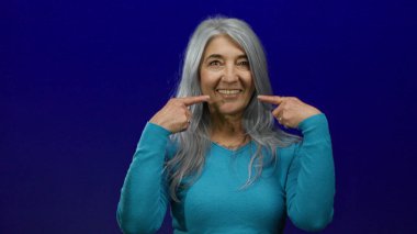Gri saçlı yaşlı bir kadın, izole edilmiş mavi bir arka plana sıcak bir şekilde gülümsüyor, rahatlatıcı hareketlerle neşeli yüzüne işaret ediyor, kendine güvenen, huzurlu bir anı yakalıyor..