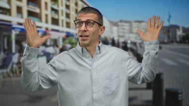 Kalabalık bir sokakta, gözlüklü ve tişörtlü, şehir binaları ve arka plandaki insanlarla çevrili bir adam şaşkınlığını ifade ediyor..