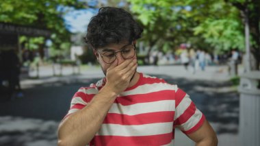 Yeşil ağaçlar ve bulanık yayalarla dolu işlek bir caddede, çizgili dışarıda dolaşan İspanyol bir adam kötü kokuya karşı burun kıvırıyor..