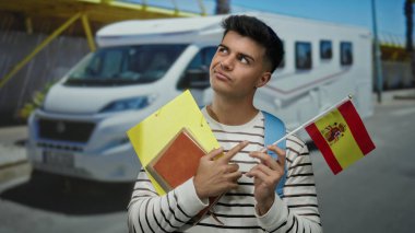 İspanyol bayrağı ve not defterleri taşıyan genç adam, şehir caddesinde park halindeki bir kampçıya karşı düşünceli bir şekilde duruyor, çizgili bir gömlek ve sırt çantası giyiyor, seyahat ve eğitimi simgeliyor..