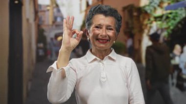 Beyaz bluz giyerek şehir caddesinde güzel jest yapan gri saçlı gülümseyen kadın, canlı şehir atmosferine pozitif ve güven yansıyor..