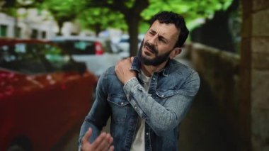 Ağaçları ve arabaları olan, kot ceketli ve sakallı, üzgün bir ifadeyle omzunu tutarak rahatsızlığını gösteren İspanyol bir adam..