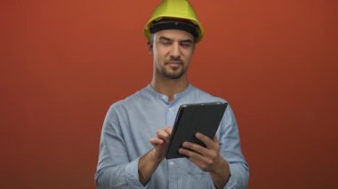 Sert şapkalı genç İspanyol adam izole edilmiş kırmızı arka plana karşı tablet kullanıyor. Bu da modern inşaat teknolojisinin odaklanmış ifadeye sahip olduğunu gösteriyor..