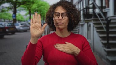 Kıvırcık saçlı ve gözlüklü bir kadın çıplak elini kaldırır ve park halindeki arabalarla dolu bir caddede kırmızı bir kazakla göğsüne avuç içini yerleştirir..