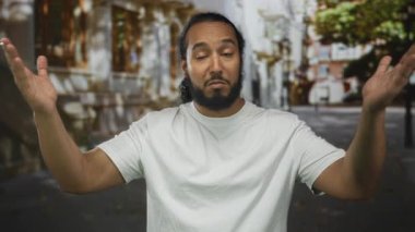 Sakallı ve kıvırcık saçlı bir adam taşın yanındaki bir caddede omuz silkip beyaz tişört giymiş ve ikna olmamış bir halde ağaçların önünde omuz silkiyor..