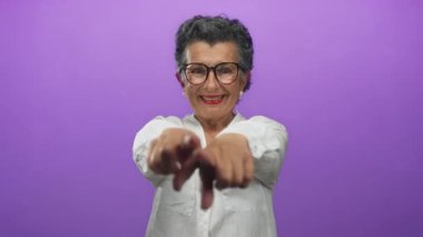 Gri saçlı ve gözlüklü gülümseyen bir kadın canlı mor bir arkaplan karşısında sevinçle el kol hareketi yapıyor beyaz bir gömlek giyiyor, pozitif yaşlanma ve neşeyi temsil ediyor..