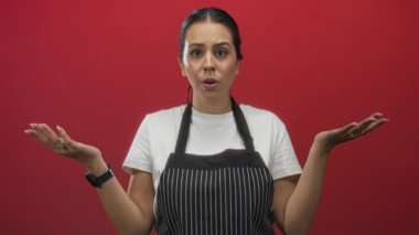 Çizgili önlük ve beyaz tişört giyen, kırmızı stüdyoda omuz silkme hareketi yapan genç bir kadın..