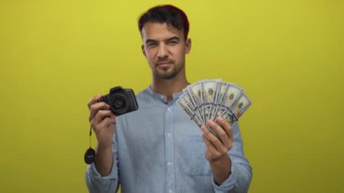 Elinde kamera olan genç bir adam bize izole edilmiş sarı arka planda dolar gösteriyor. Bu da fotoğrafçılıkta ve hareketli bir ortamda finansal kazanç sağladığını gösteriyor..