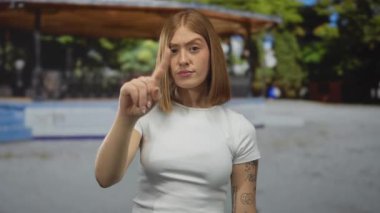 Genç sarışın kadın, güneş ışığında parkın sağını işaret ediyor; talimat uyarı odaklı kararlılık.