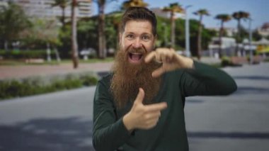 Uzun sakallı bir adam, gün ışığında bir şehir caddesinde gülümserken parmaklarıyla bir sahne çerçeveler; heyecan yaratıcılık.