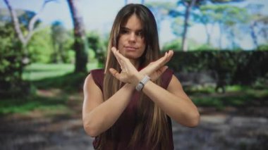 Yeşil bir parkta dikilen İspanyol bir kadın reddedilme jesti olarak kollarıyla X oluşturarak sakin bir doğal ortamda hoşnutsuzluğunu dile getiriyor..