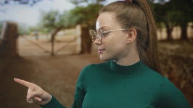 Yuvarlak gözlük takan ve yeşil kazak giyen genç beyaz kadın ormanı işaret ediyor; keşif yönü merakı.