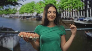 Amsterdam caddesinde kanalın kenarında dururken elinde yumurta tepsisi ve kıvırcık saç tutan İspanyol genç kadın..
