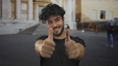 Sakallı genç adam güneşli bir günde arka planda tarihi mimarisi olan şehir ortamında mutluluk ve pozitiflik saçan dışarıda başparmak kaldırıyor..