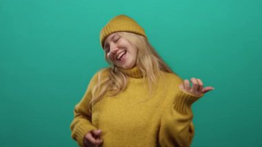 Sarı kazak giyen ve bere takan bir kadın canlı yeşil arka planda gitar çalarken neşeli, gençlik enerjisi ve ifadesini yansıtıyor..