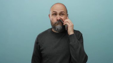 Telefonda konuşan kel bir adam mavi arka planda hareketler yapıyor. İletişim kurarken kafa karışıklığı ve hayal kırıklığı gösteriyor.