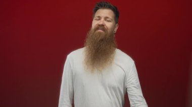 Kızıl stüdyoda gülümseyen ve parmağıyla işaret eden uzun sakallı beyaz bir genç..