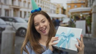 Parti şapkası takan, gülümseyen ve elinde mavi kurdeleli bir hediye kutusu olan bir kadın. Mutluluk kutlaması..