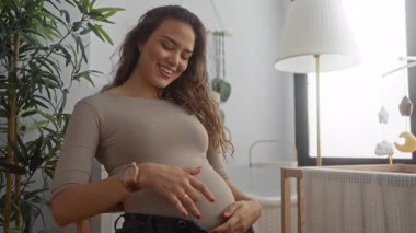 Bebek mobilyası ve bitkilerle çevrili rahat bir yatak odasında karnına hafifçe dokunarak gülümseyen hamile kadın annelik için neşe ve beklenti saçıyor..