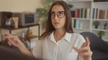 Gözlüklü ve beyaz tişörtlü genç bir kadın kitaplığın ve kahve istasyonunun yanındaki ofis masasında gözleri kapalı bir şekilde umut dolu bir dilek diliyor..