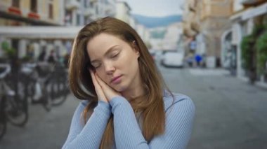 Mavi kazaklı genç beyaz kadın kalabalık bir caddede elleri yanağına bastırır; dinlenme huzur içinde uyu.