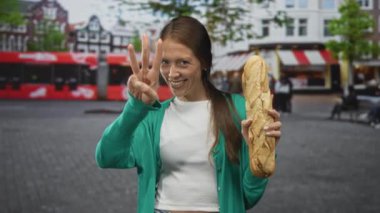 Genç İspanyol kadın bir baget tutuyor ve gülümserken üç parmağını gösteriyor, yeşil hırka ve beyaz bluz giyiyor; neşe topluluğu.