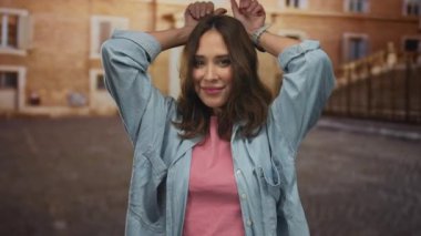 Günlük kot ve pembe elbiseli genç İspanyol kadın hareketli bir şehir caddesinde eğlenceli bir jest yapıyor, meşgul bir şehir ortamında neşe ve sıcaklık saçıyor..