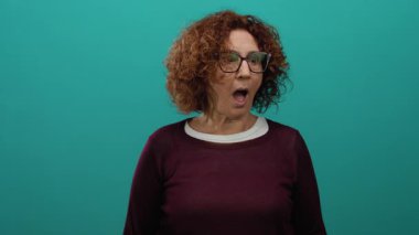 Kıvırcık saçlı ve gözlüklü bir kadın canlı yeşil bir arka plana karşı şaşkınlığını dile getiriyor, açık ağzıyla duyguları çağrıştırıyor ve gündelik, modern bir tarz sergiliyor..