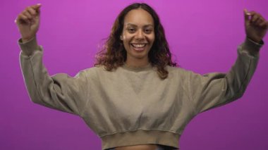 Mor bir stüdyoda, iki işaret parmağıyla gülümseyen ve işaret eden genç Afrikalı Amerikalı kadın..
