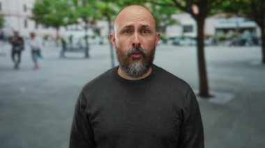 Bulanık yayalar ve yeşilliklerle çevrili, hareketli bir şehir sokağında açık havada sürpriz yapan kel bir adam, samimi bir şehir havası yakalıyor..
