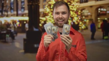 Turuncu mahkum tulumlu bir adam, elinde bir yığın parayla, elinde tatil ışıkları ve ağacı olan bir sokakta, bayram yaramazlığı yapan bir adam..