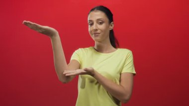 Kırmızı stüdyoda sarı tişört giyerken açık avuçlar sunan gülümseyen genç İspanyol kadın; ürünün neşeli görüntüsü.