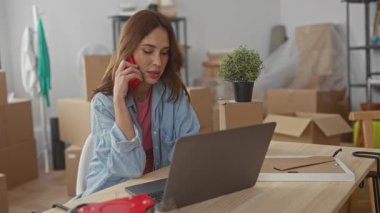 Yeni dairede telefonla konuşan bir kadın, paketleri açılmamış kutularla iç dizüstü bilgisayarda çalışıyor. Etrafı bitkiler ve dekorlarla çevrili. Yeni taşındığını gösteriyor..