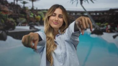 Gülümseyen sarışın kadın, tropikal havuz başındaki palmiye ağaçlarını işaret ediyor; dostça bir davet.
