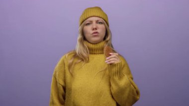 Sarı kazak giyen ve şapkalı bir kadın mor arka planda kurabiye tutuyor. Parmak hareketleriyle onaylamadığını gösteriyor. Anlaşmazlık ya da inkar olduğunu gösteriyor.