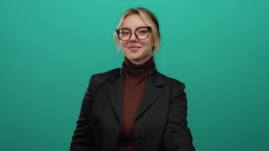 Kadın kalp hareketi yapıyor, parmakları yeşil bir zemine gülümsüyor, sarı saçları ve gözlükleriyle kendine güveni ve pozitifliğini gösteriyor..