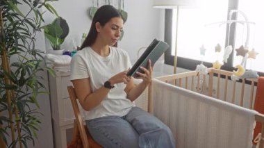 Yatak odasındaki sandalyeye oturmuş, tahta bebek beşiği ve asılı mobil yanında tablet ekranı tıklatan kadın, sakin dikkat ebeveynlik.