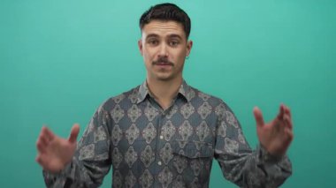 Genç İspanyol adam yeşil arka plan duvarının önünde elleri ile büyük jest yapıyor, desenli gömlek ve küpeler giyiyor, boyunu veya mesafesini ifade ediyor..